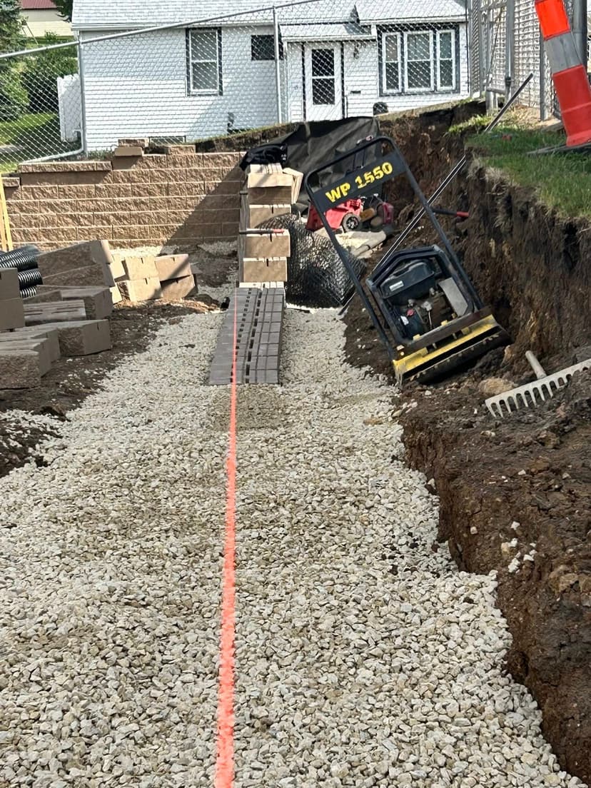 Construction site featuring excavated trench, gravel path, and equipment for landscaping installation.